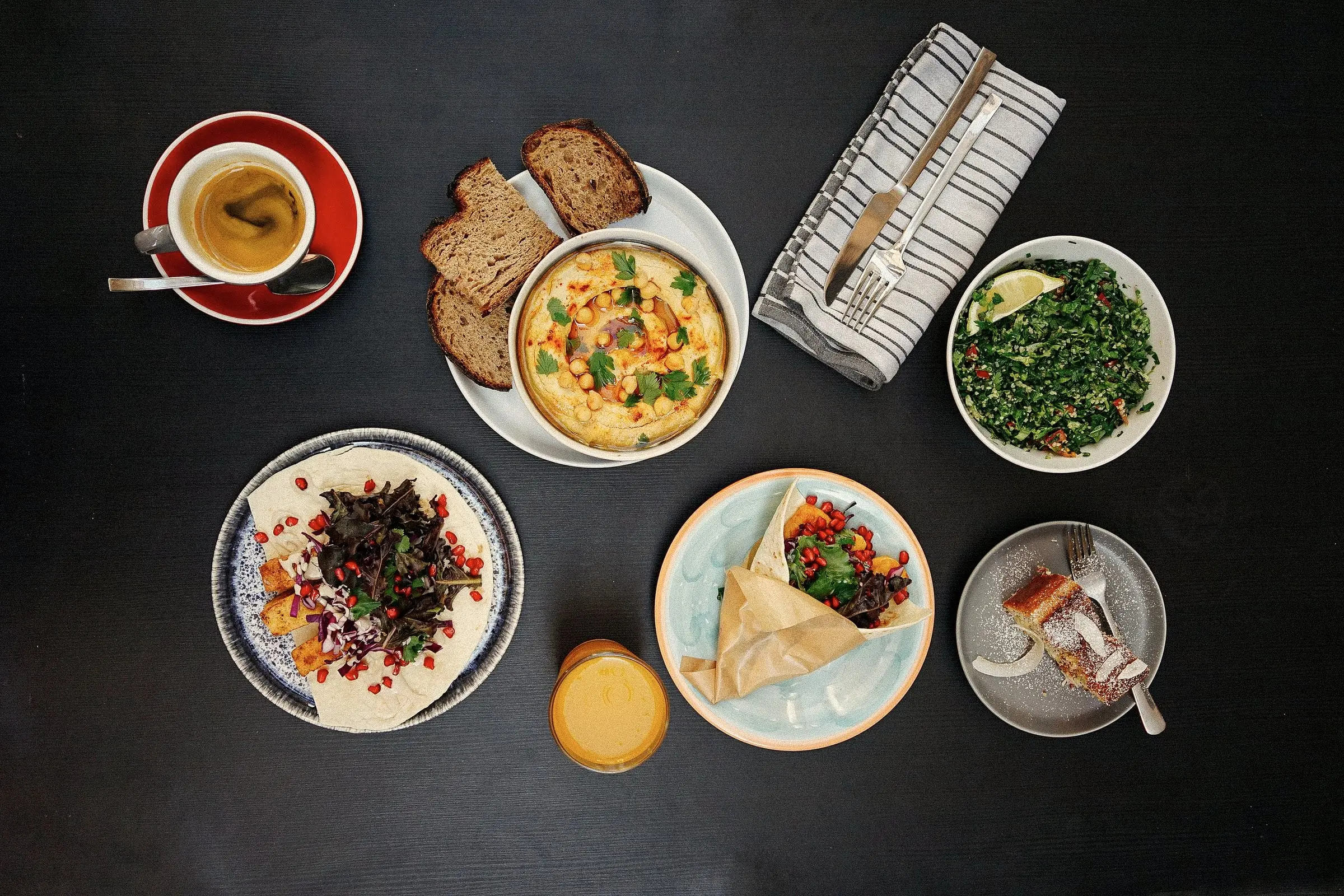Brunchrätter sedda uppifrån på ett mörkt bord: hummus, bröd, sallad och kaffe.